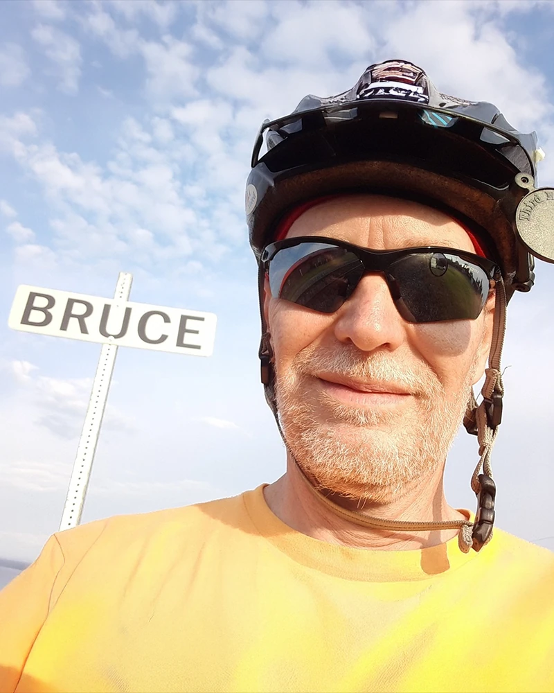 Bruce Sarjeant wearing a bicycle helmet and sunglasses next to a street sign that reads Bruce.