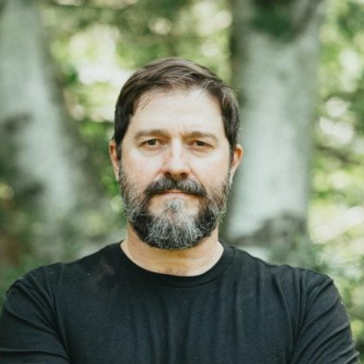 M. Bartley Seigel with a beard and black t-shirt outdoors against a blurred forest background.
