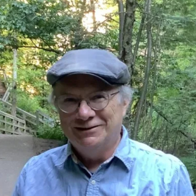 Russell Thorburn wearing glasses and a flat cap outdoors in a wooded park setting.