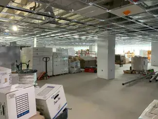 Stacks of construction supplies on pallets, exposed ceiling grid, and a drywall-covered support pillar.